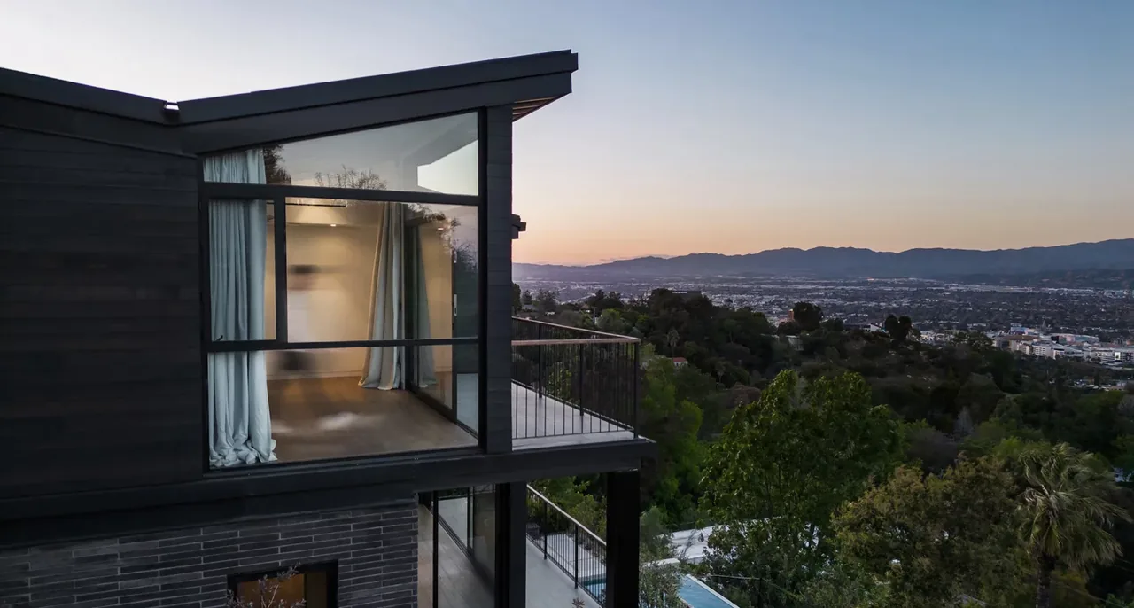 Exterior view of a modern two-story house with large glass windows and a balcony overlooking a hilly cityscape at sunset.