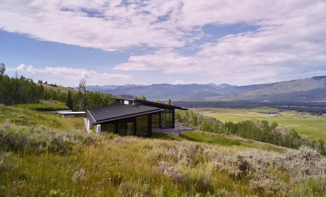 Modern house with sloping black roofs set in grassy hills with mountains in the background under a partly cloudy sky.