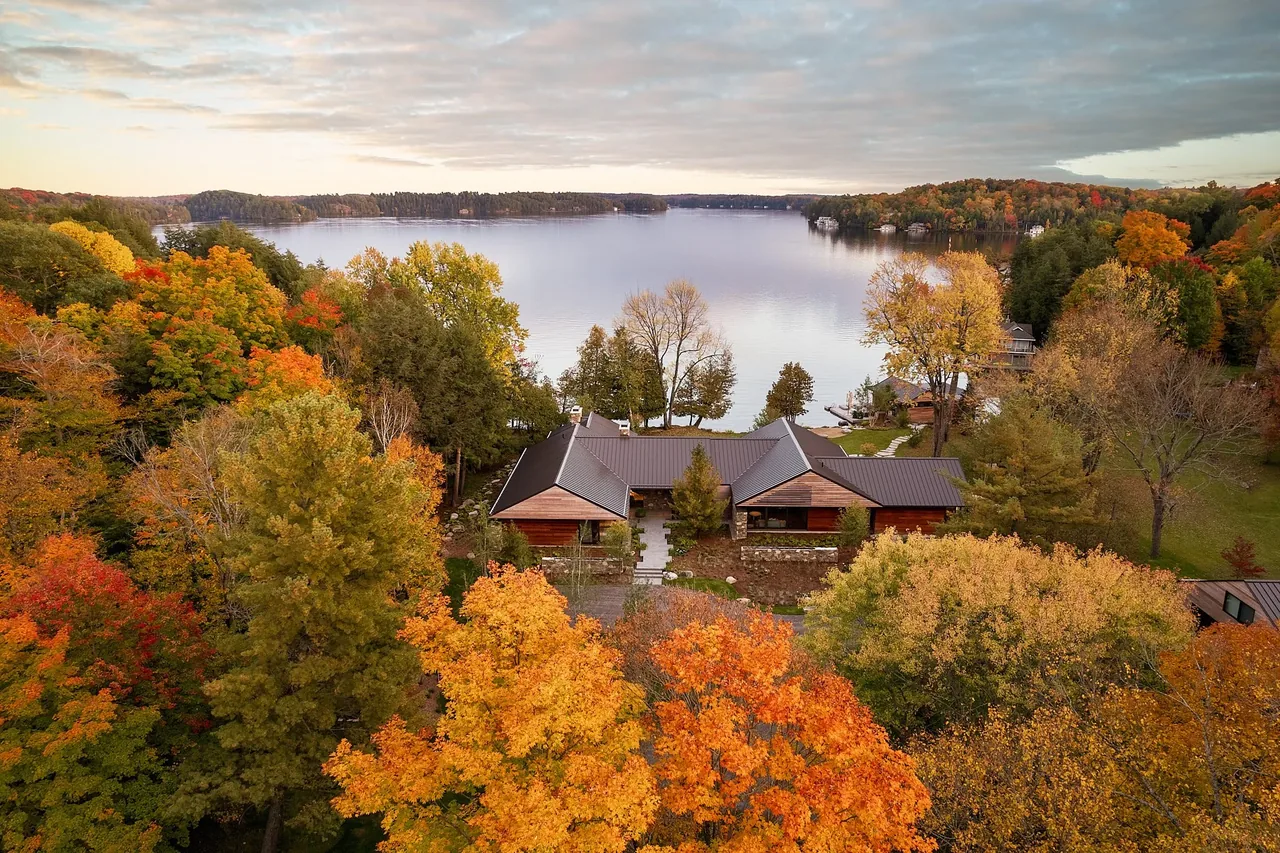 Aerial view of a cottage surrounded by vibrant fall trees with a lake in the background.