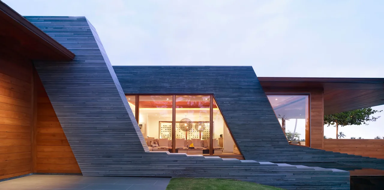 Exterior front view of a modern residence featuring angled dark wood cladding and large floor-to-ceiling windows revealing the lit interior.