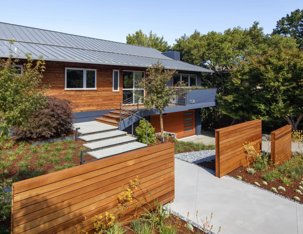 Modern suburban house with wooden siding, flat metal roof, and a landscaped front yard with a wooden fence and concrete steps.
