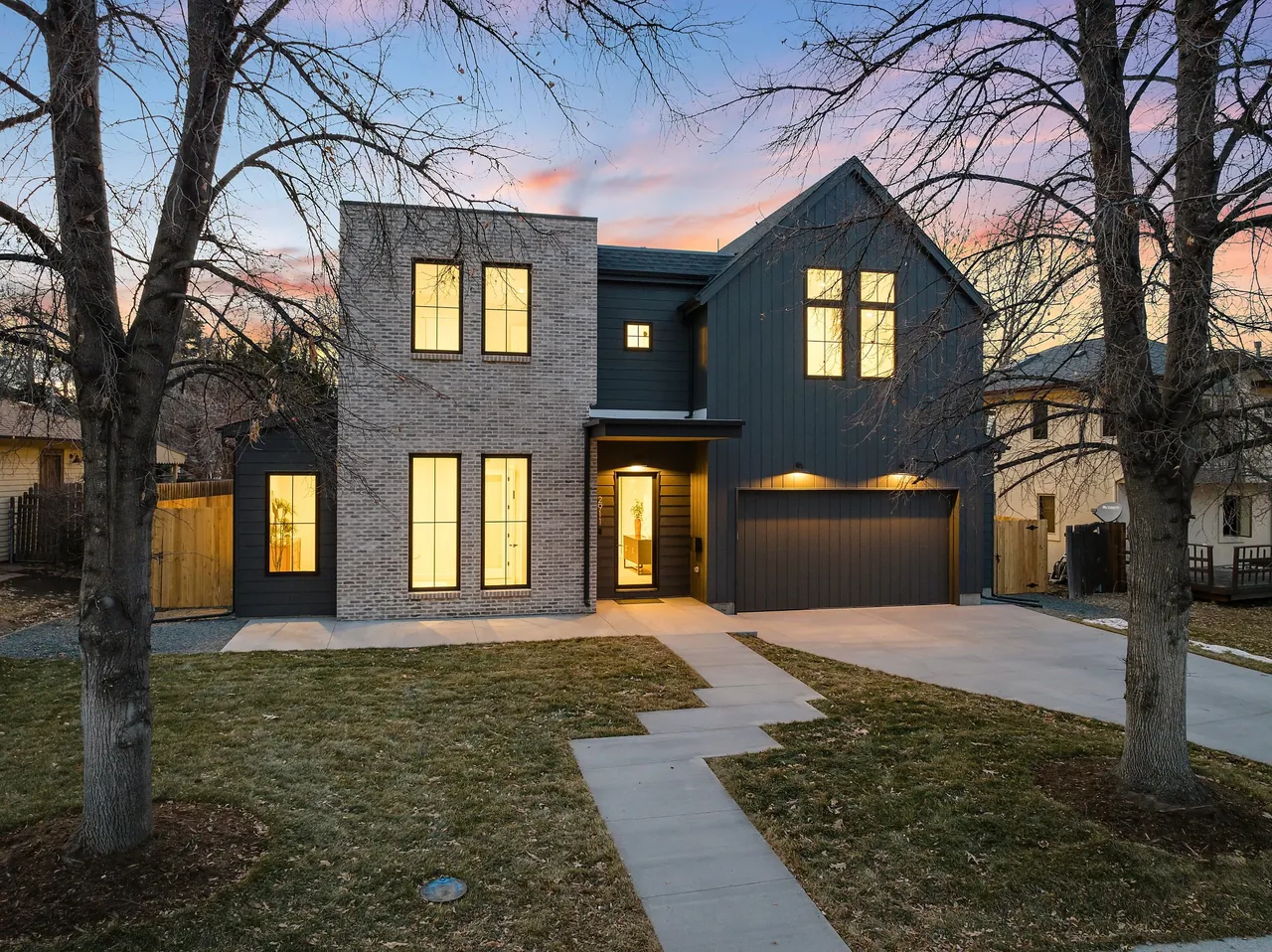 Two-story residential house with brick and dark siding exterior at dusk with lit windows and driveway.