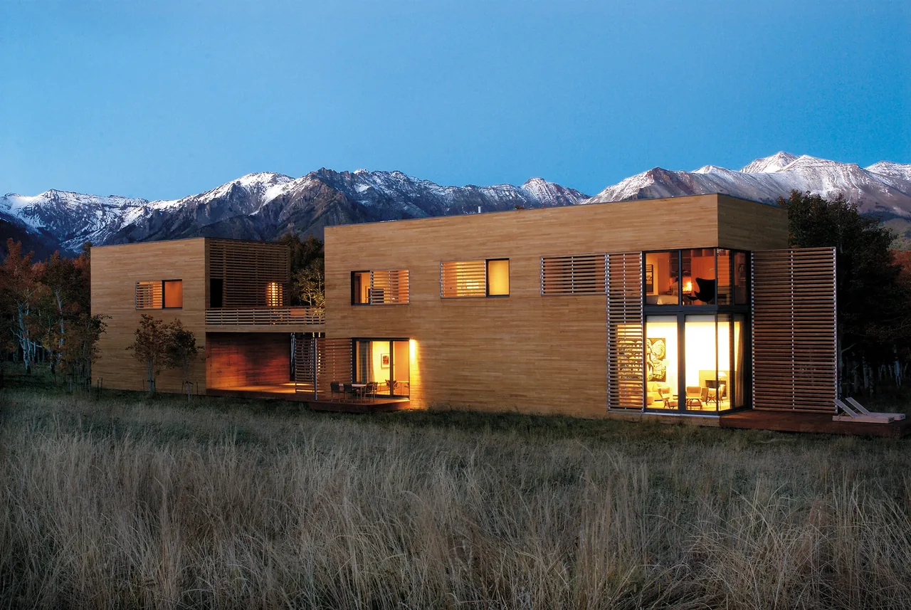 Two-story modern wooden house with horizontal slats and large windows at dusk, set against a backdrop of snow-capped mountains and grassy me