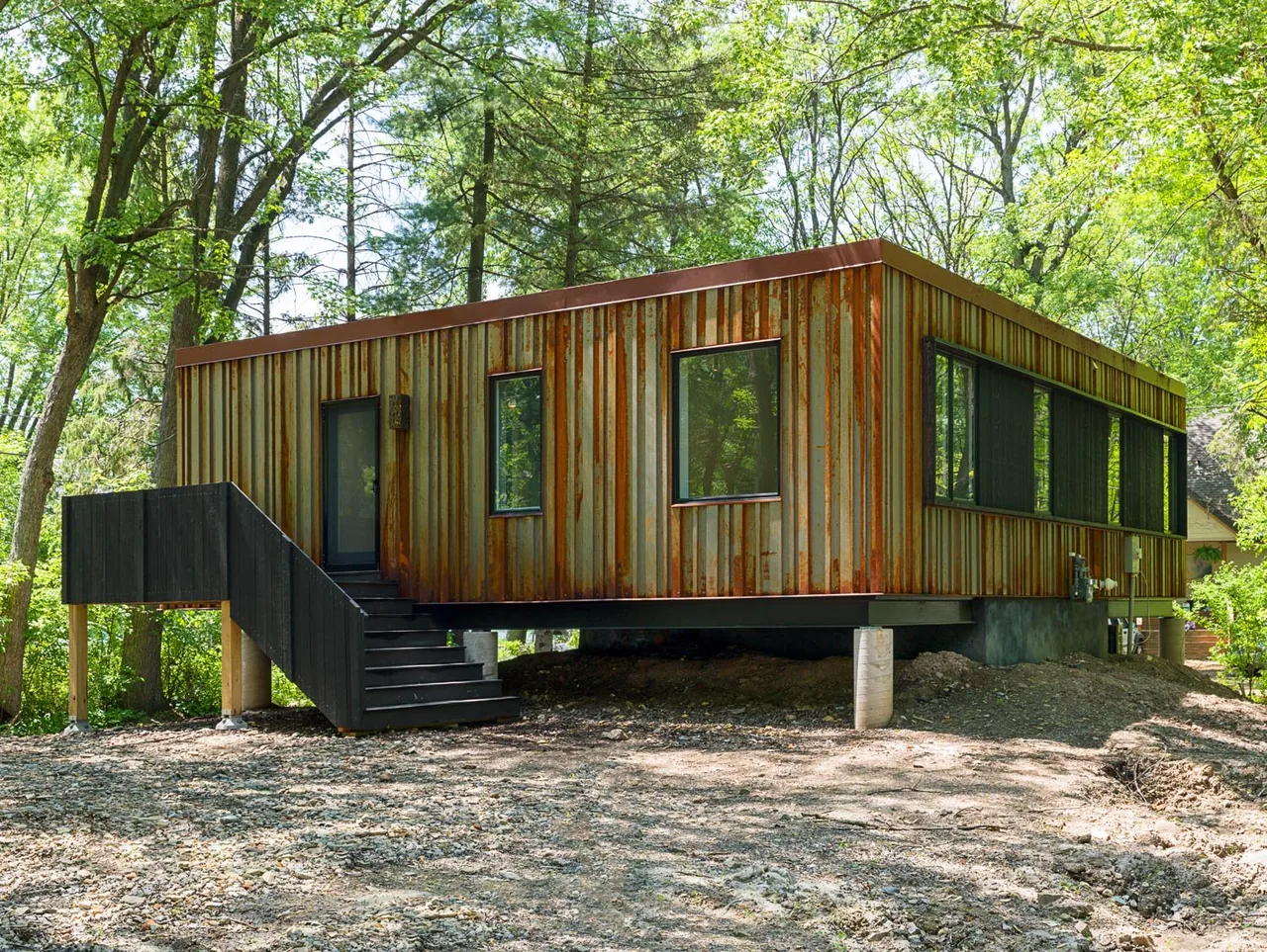 Modern elevated rectangular house with rusted metal siding and black framed windows surrounded by trees.