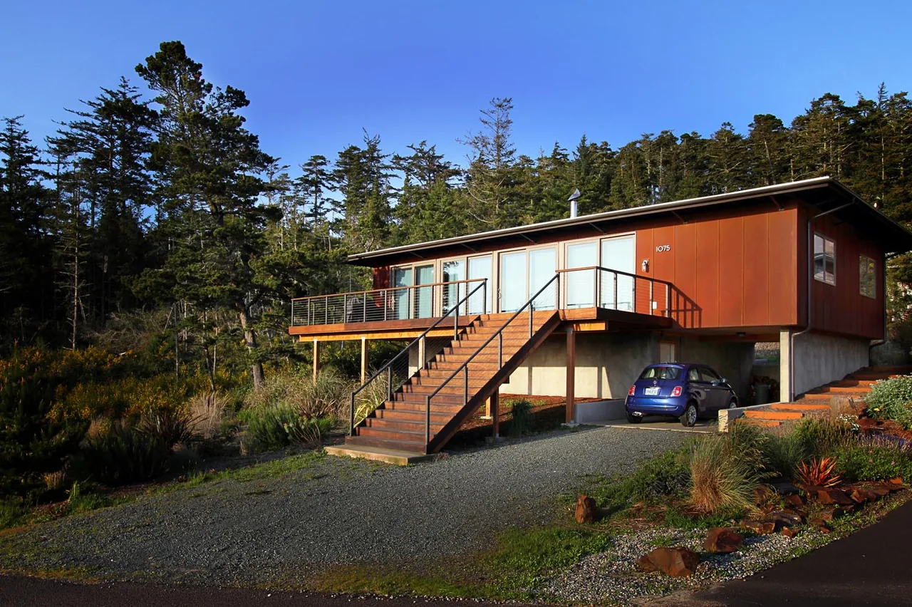 Modern elevated house with rust-colored exterior panels, wooden stairs and deck with cable railing, surrounded by trees and landscape.
