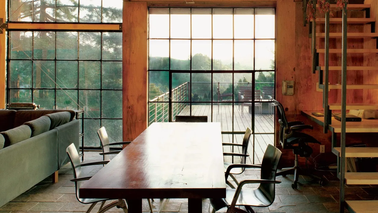 Interior view of a dining and office area with large metal-framed windows overlooking an outdoor deck with seating.