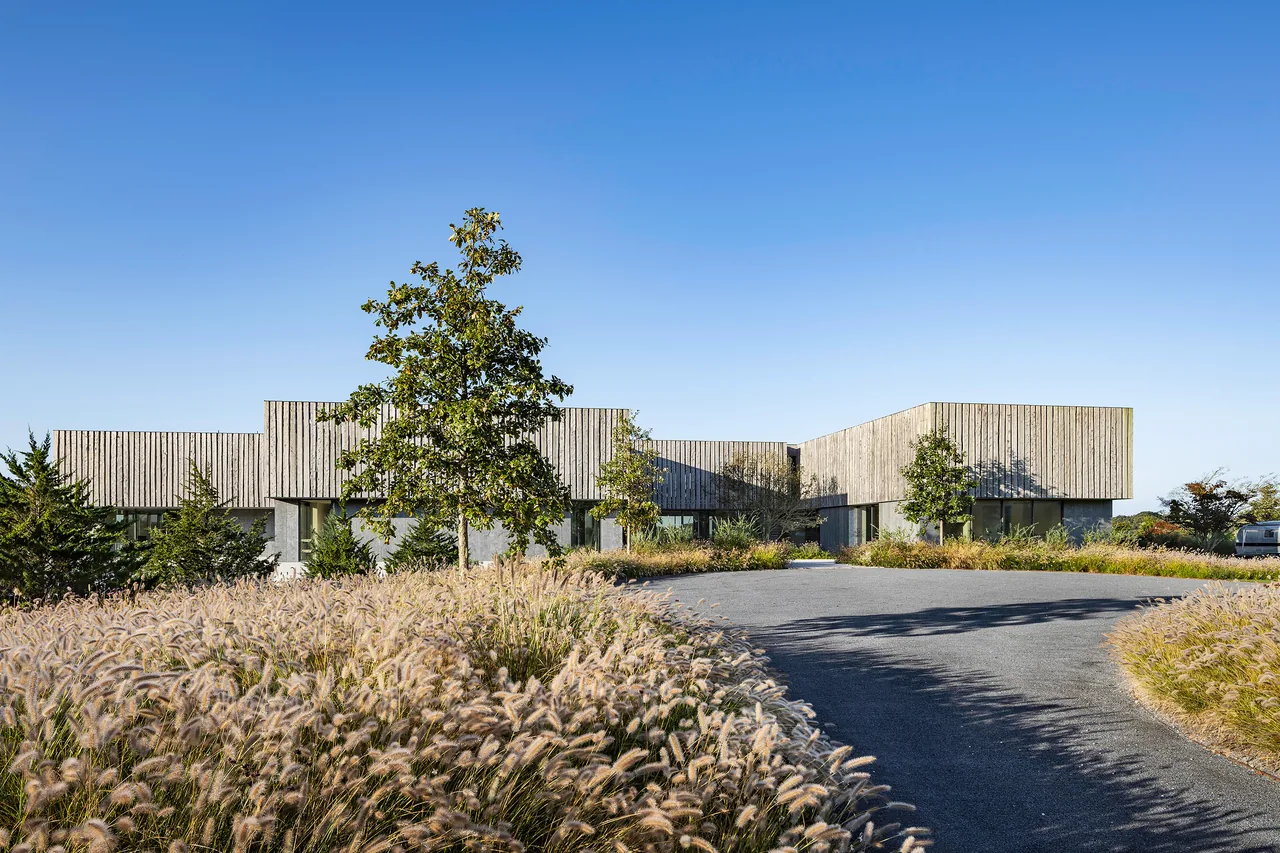 Single-story modern building with a façade of vertical wood cladding, surrounded by landscaped grasses and trees under a clear sky.