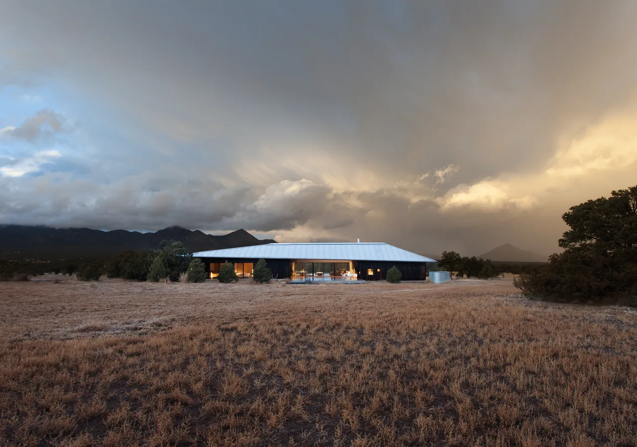 Single-story ranch house with a metal roof set in dry grassland under dramatic cloudy sky.