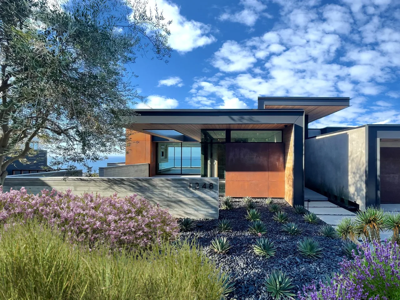 Modern single-story house with flat roofs, large windows, and mixed materials including concrete and rust-colored panels, surrounded by succ
