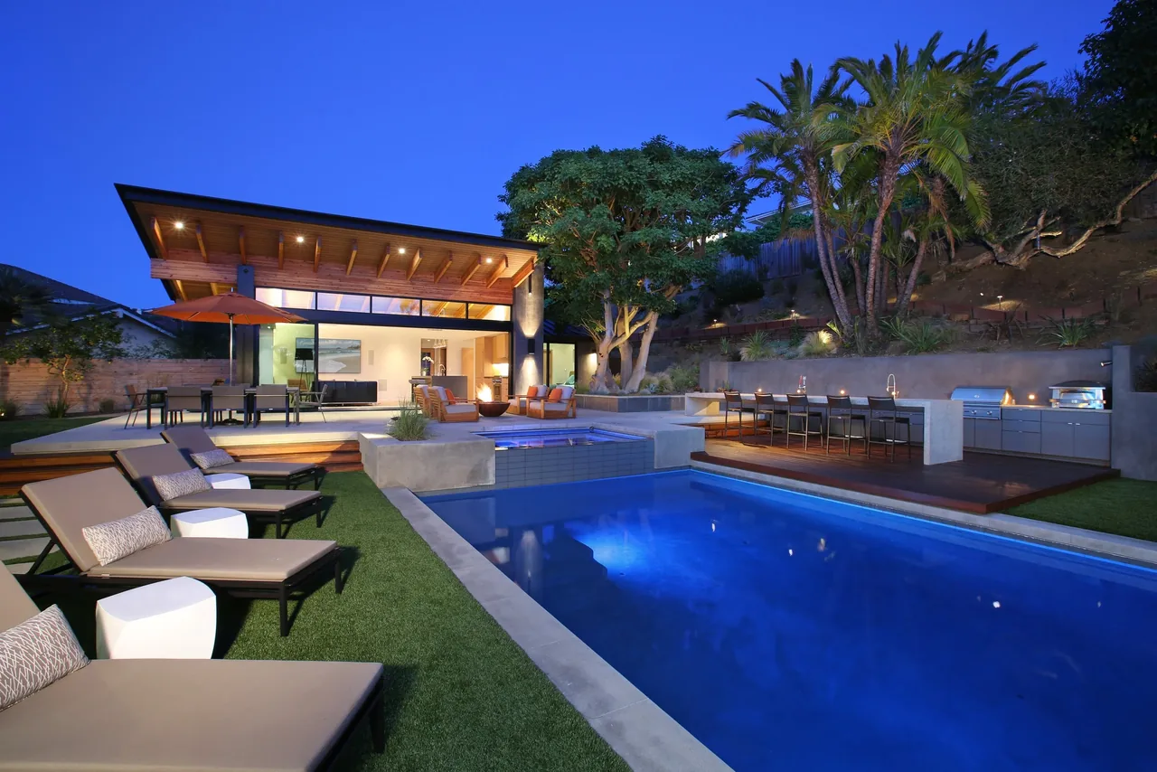 View of a modern pool house at twilight featuring an illuminated swimming pool, lounge chairs, and an outdoor kitchen area.