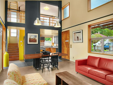 Interior view of a living and dining area with large windows, colorful furniture, and a kitchen in the background.
