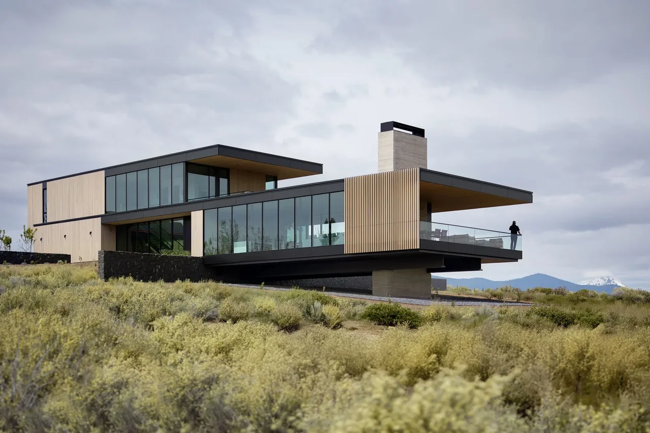 Modern two-story house with extensive glass walls, cantilevered deck, and wood siding situated in a natural landscape.