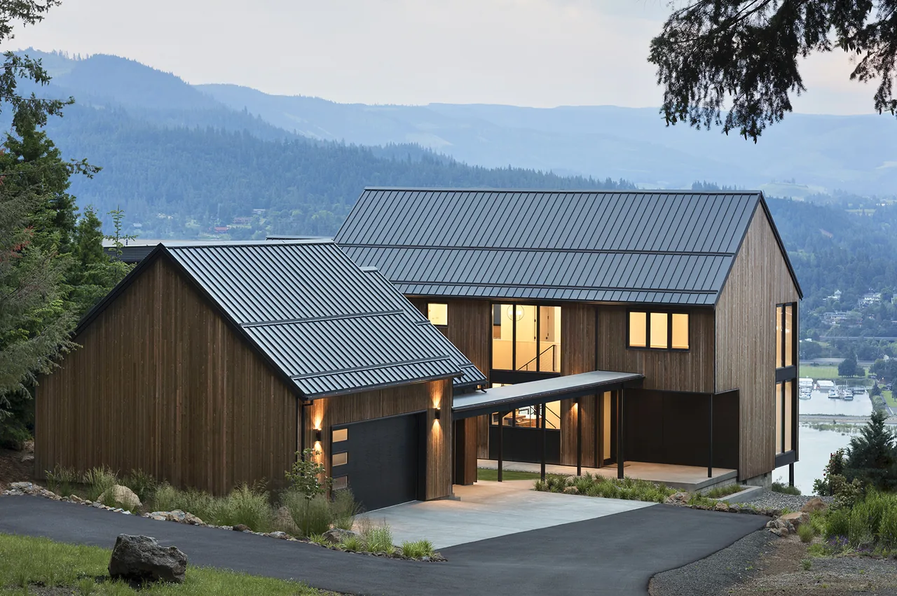Modern house on a hilltop with dark metal standing seam roofs and wood siding overlooking a valley and water body at dusk.