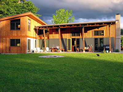 Exterior view of a two-story wooden house with large windows and a covered patio under a partly cloudy sky.
