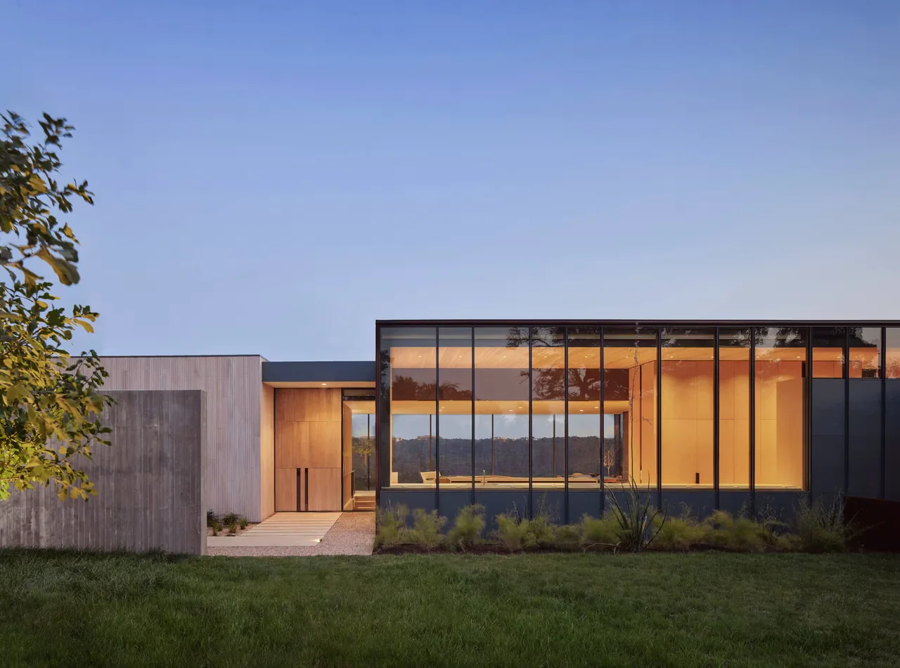 Exterior view of a modern one-story house with floor-to-ceiling glass windows and wood accents at dusk.