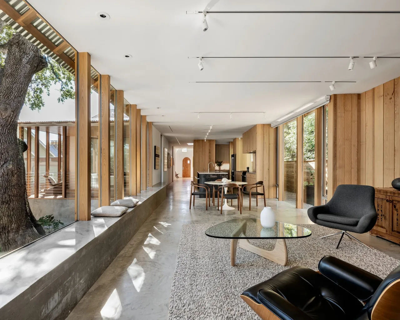 Long open living and dining area with concrete floors, wood panel walls, and large floor-to-ceiling windows overlooking an outdoor deck and,