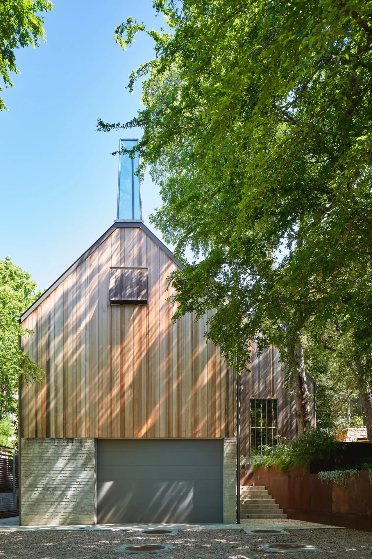 Front view of a modern house with wood siding, concrete base, and a tall vertical glass chimney-like structure, surrounded by large green-le