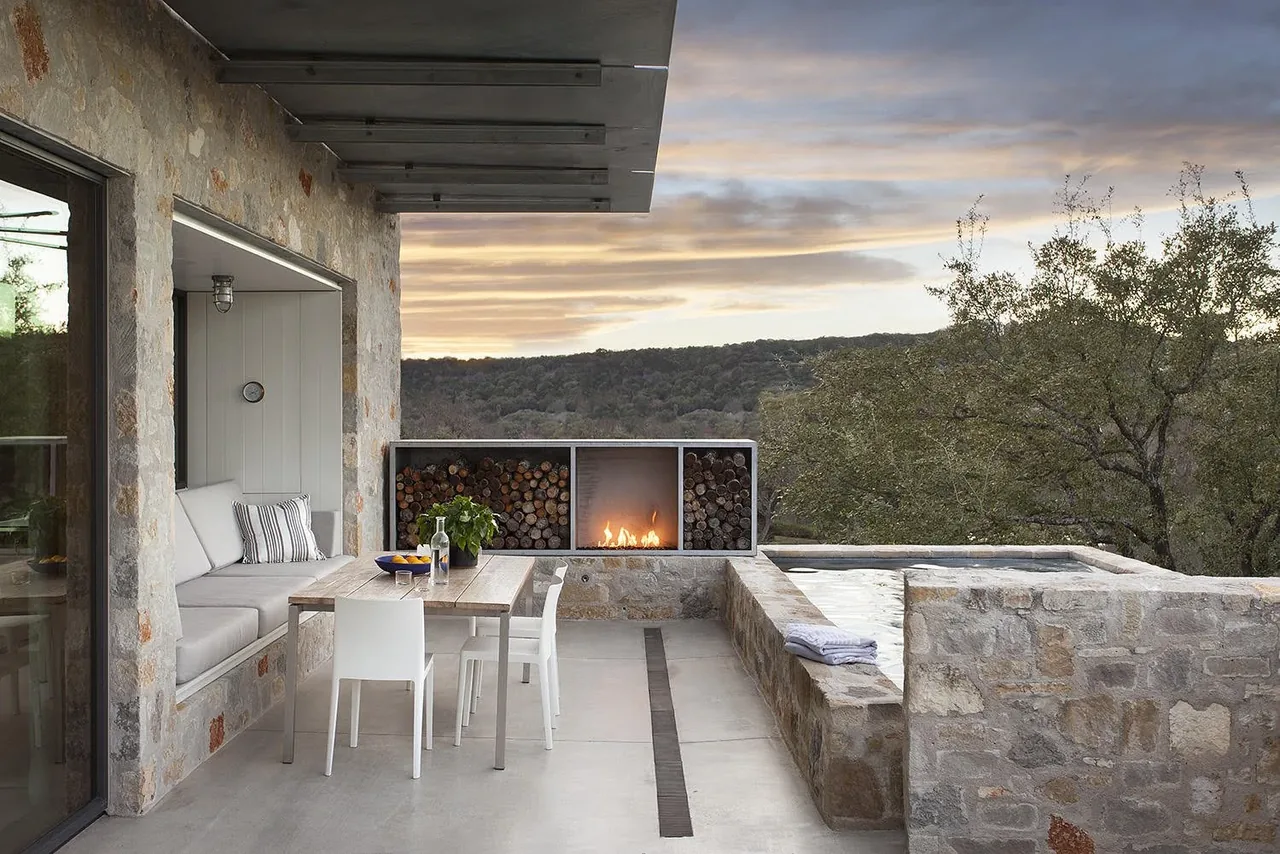 Outdoor patio with stone walls, built-in seating, dining table, modern fireplace, and a hot tub overlooking wooded hills at sunset.