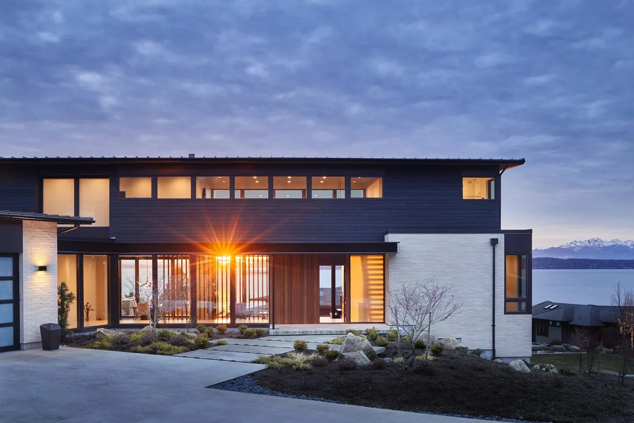 Modern two-story home exterior at dusk with large windows and black wood siding beside a white brick section.