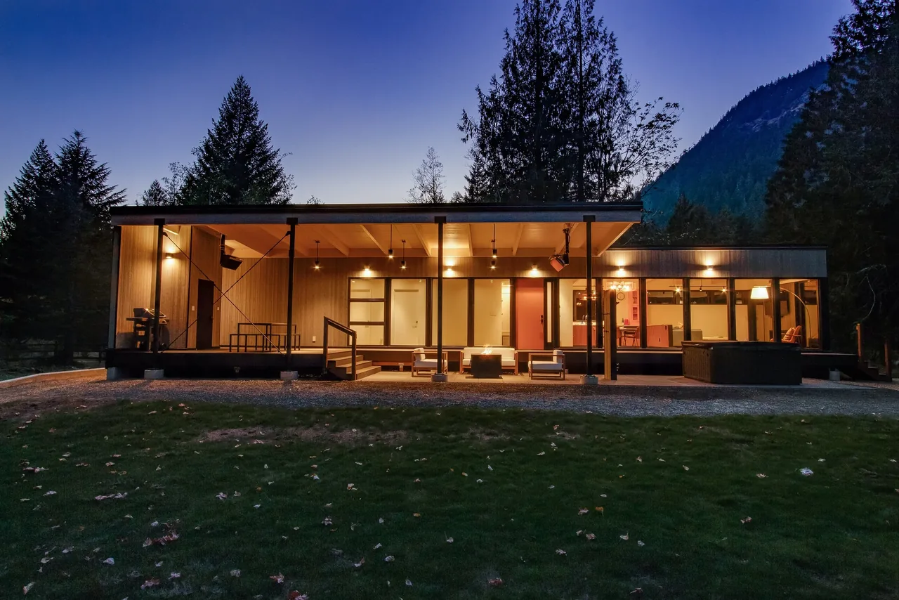 Night view of a modern cabin with wood siding and large windows in a forested mountain setting.