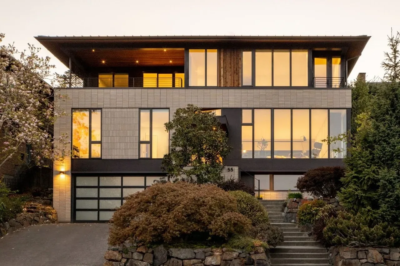 Modern multi-story house with large windows illuminated at dusk, stone landscaping, and a tree-lined driveway.