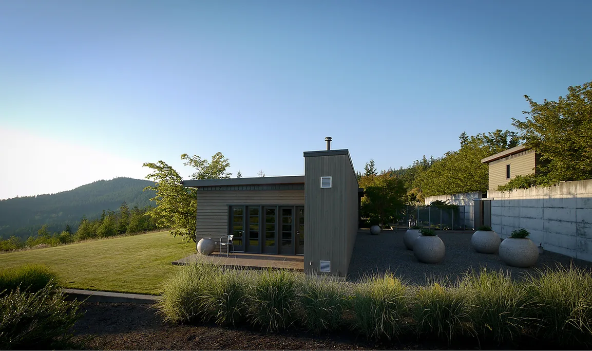 Exterior view of a modern, single-story house with a flat roof and gray wood siding, set in a landscaped garden with large round planters.
