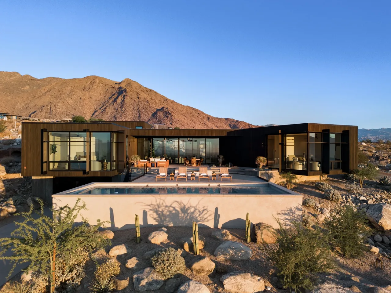 Modern single-story house with dark wood exterior and large floor-to-ceiling windows overlooking a pool in a desert landscape.