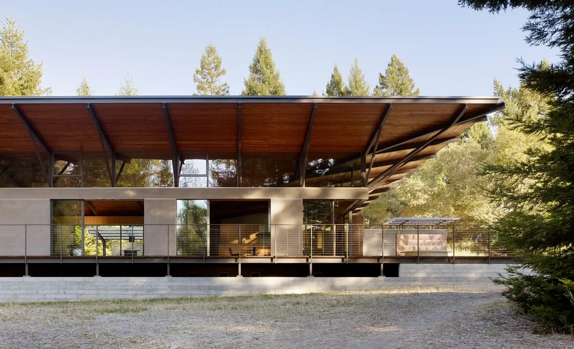 Modern house exterior with large overhanging wood roof and floor-to-ceiling glass windows surrounded by trees.