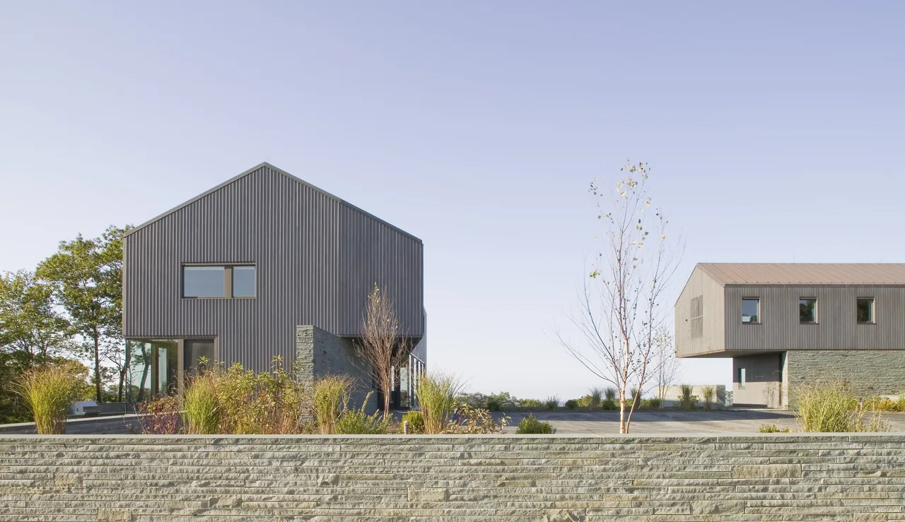 View of two modern buildings with vertical siding and stone bases separated by a paved courtyard and landscaping under a clear sky.