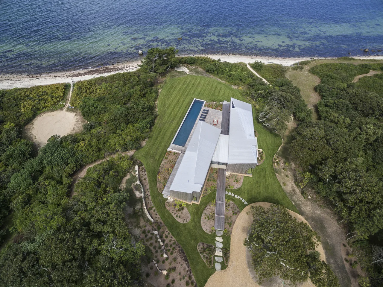 Aerial view of a modern two-shed house with a pool on a grassy lot near the water's edge.