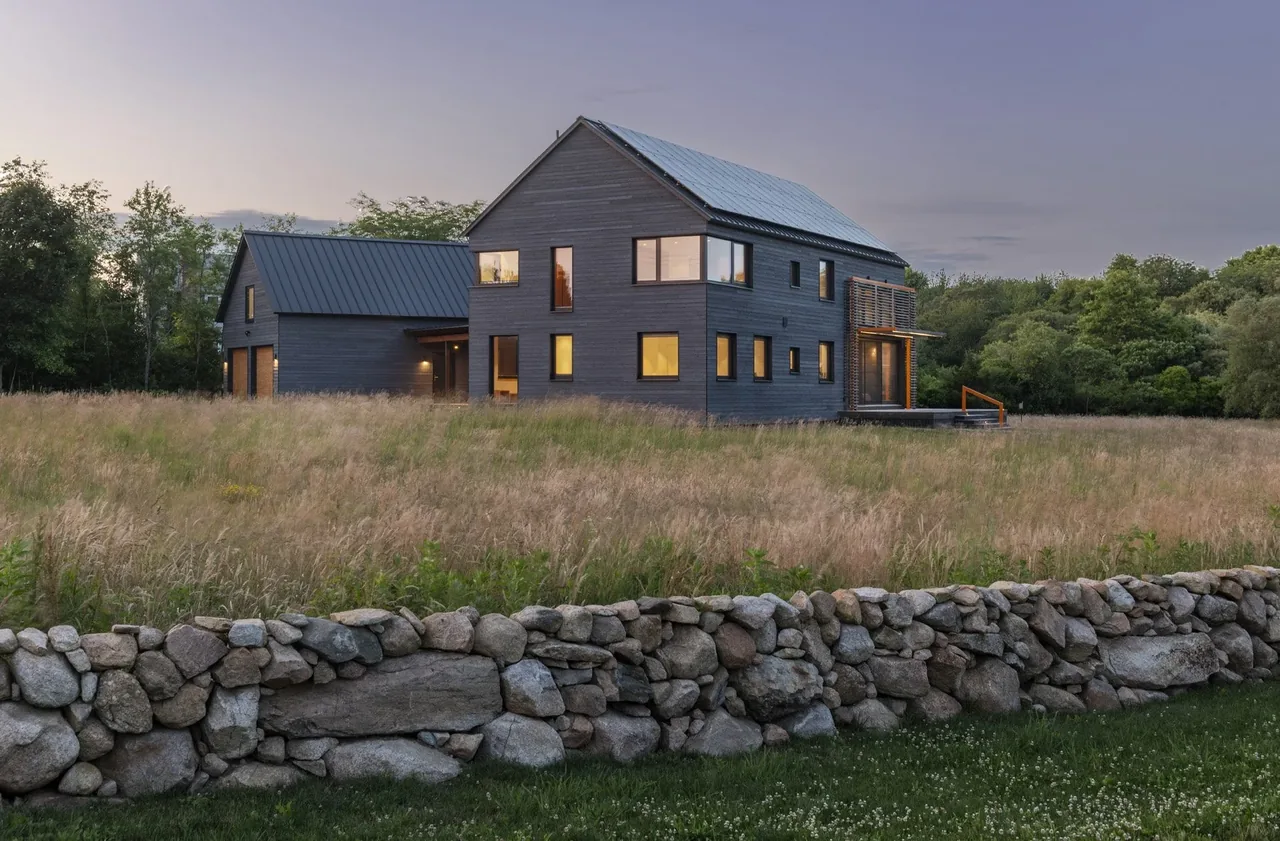 A modern two-story home with dark wood siding and metal roofs viewed across a stone wall and tall grass field at dusk.