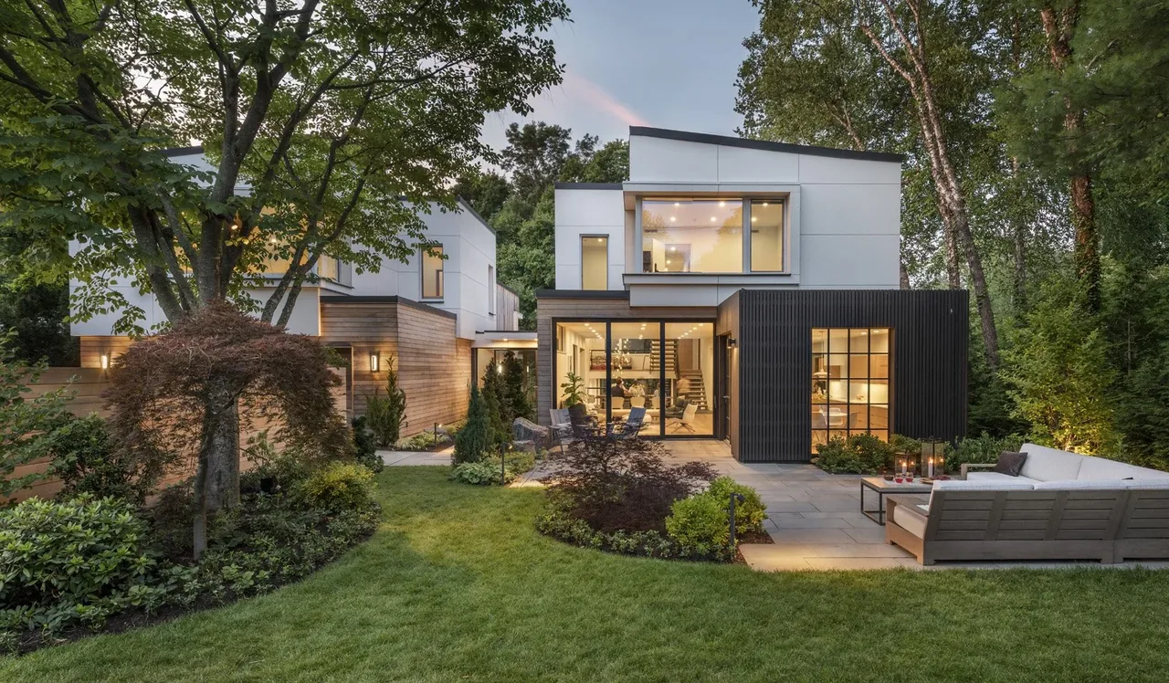 A modern two-story home with a white and dark gray exterior and large windows, surrounded by trees and a landscaped lawn at dusk.