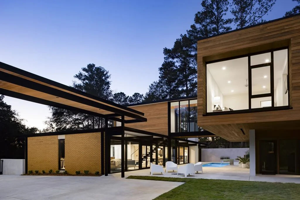 Modern two-story house with wood siding, large black-framed windows, and a partially covered outdoor seating area near a pool at dusk.