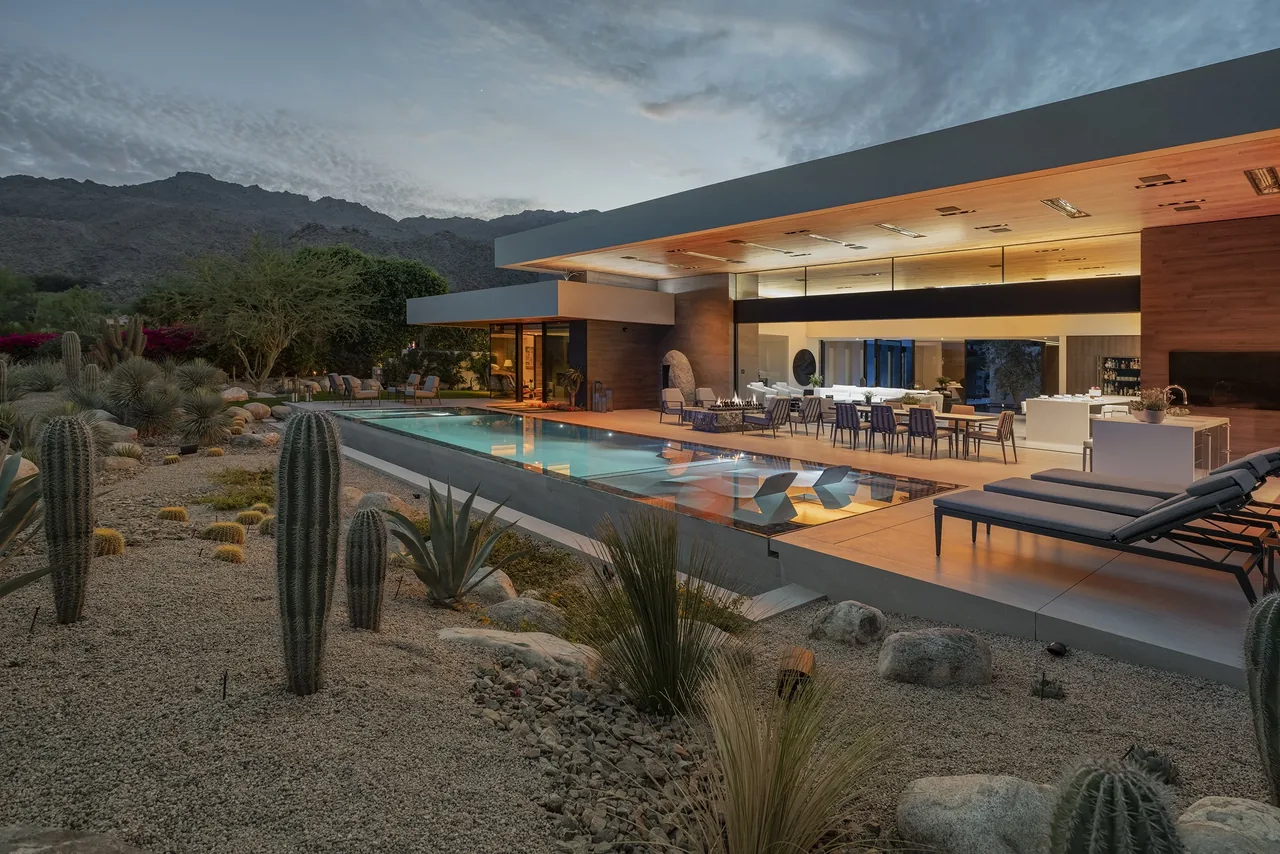 Modern home exterior at dusk featuring an open patio with pool and desert landscaping with mountains in the background.