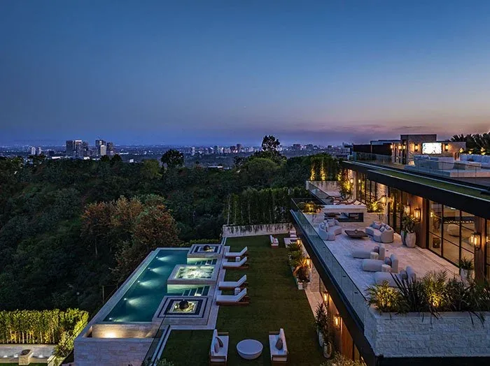 Night view of a modern terraced hillside mansion with multiple outdoor terraces, seating, and a pool overlooking a city skyline.