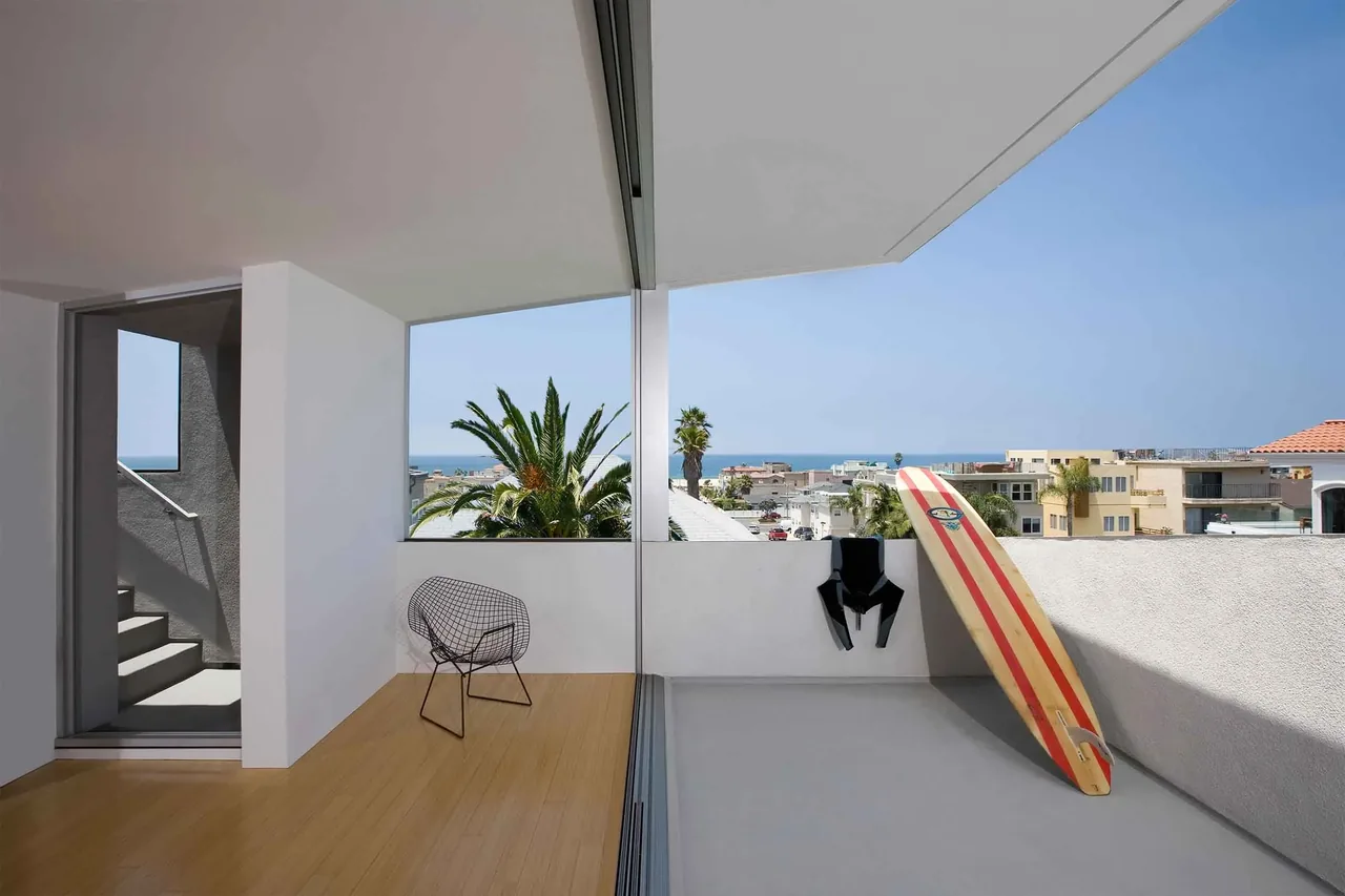 Minimalist balcony with a wire chair, surfboard, and ocean view in the distance under a clear sky.