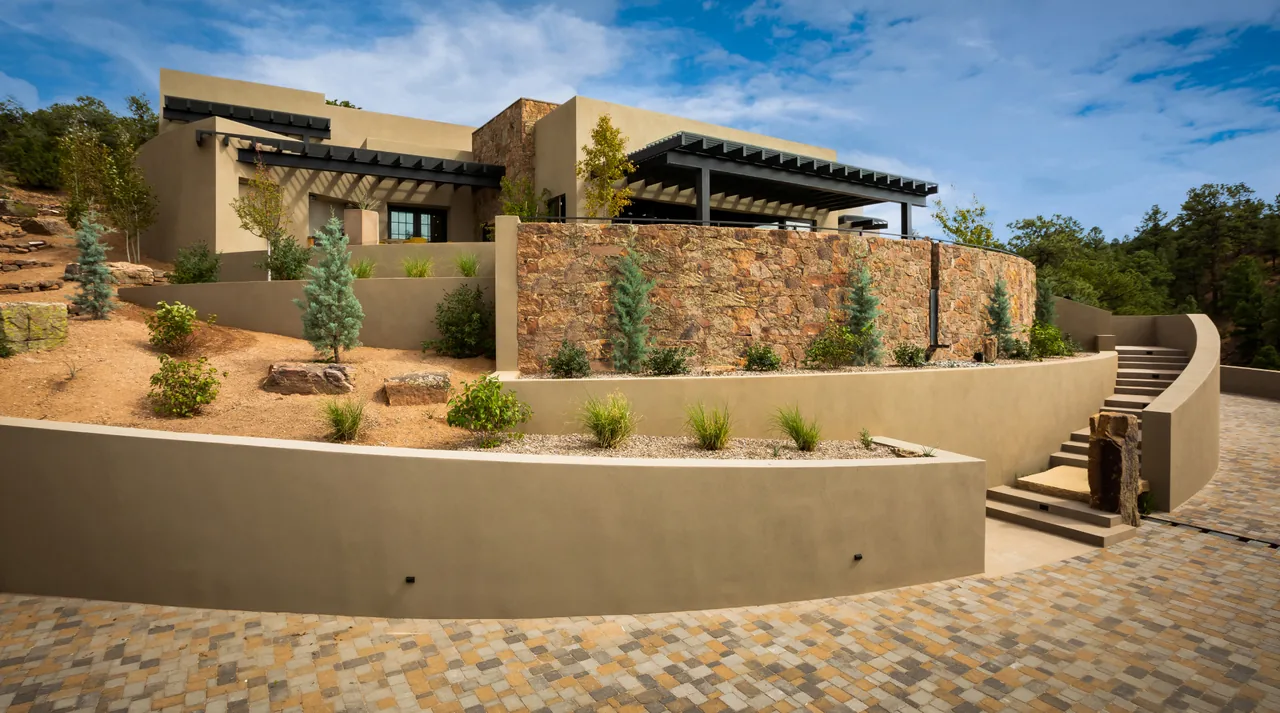 Modern house exterior with curved stone walls and a black pergola on a sunny day.