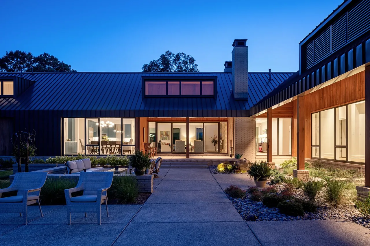 Night view of a modern home exterior with large glass doors, black metal roof, and an outdoor seating area.