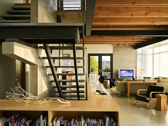 Interior view of a modern living room with exposed wooden ceiling beams, concrete walls, and a black metal staircase.