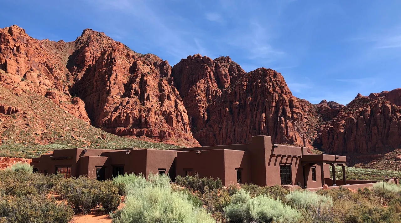 Flat-roofed adobe-style residence in a desert landscape with red rock cliffs in the background under a blue sky.