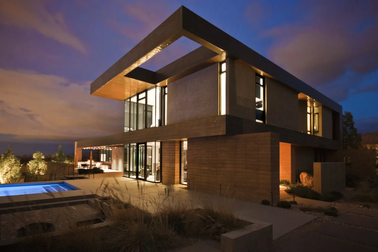 Modern two-story house at dusk with large cantilevered roof and illuminated pool in front yard.