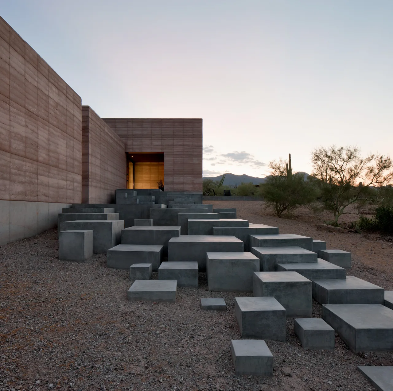 Concrete block steps lead up to a rammed earth wall entrance at dusk in a desert setting.