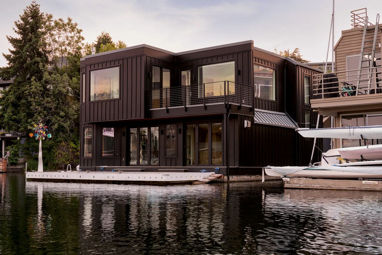 Two-story dark brown floating home with large windows and a balcony on a calm water canal.