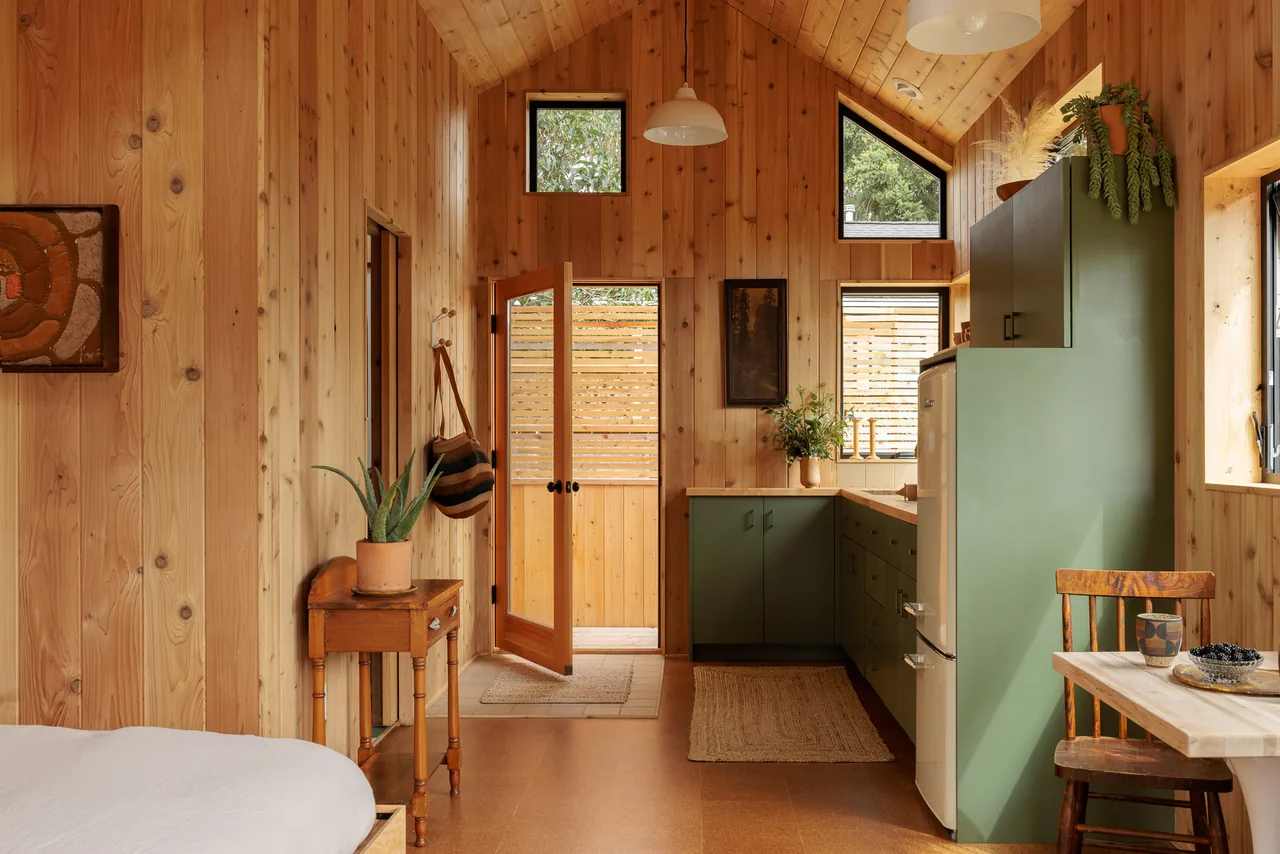 Interior of a small wooden cabin with wood-paneled walls and ceiling, green kitchen cabinets, and a wooden table with chairs.