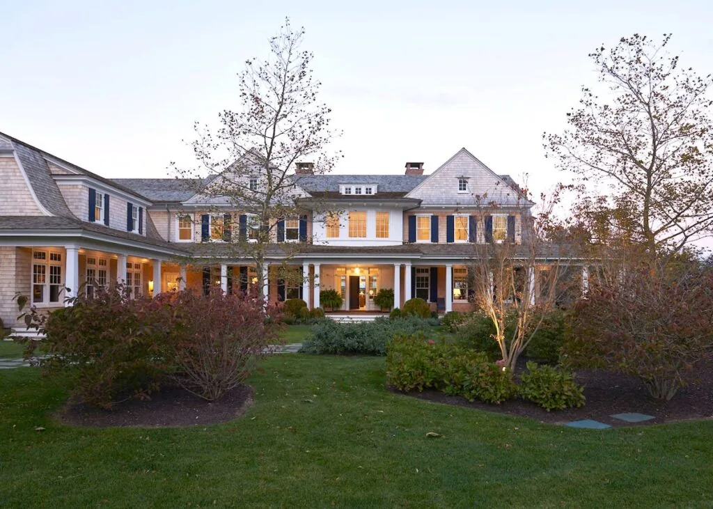 Large two-story classic American house with shingle siding, white trim, and a well-kept garden in front at dusk.