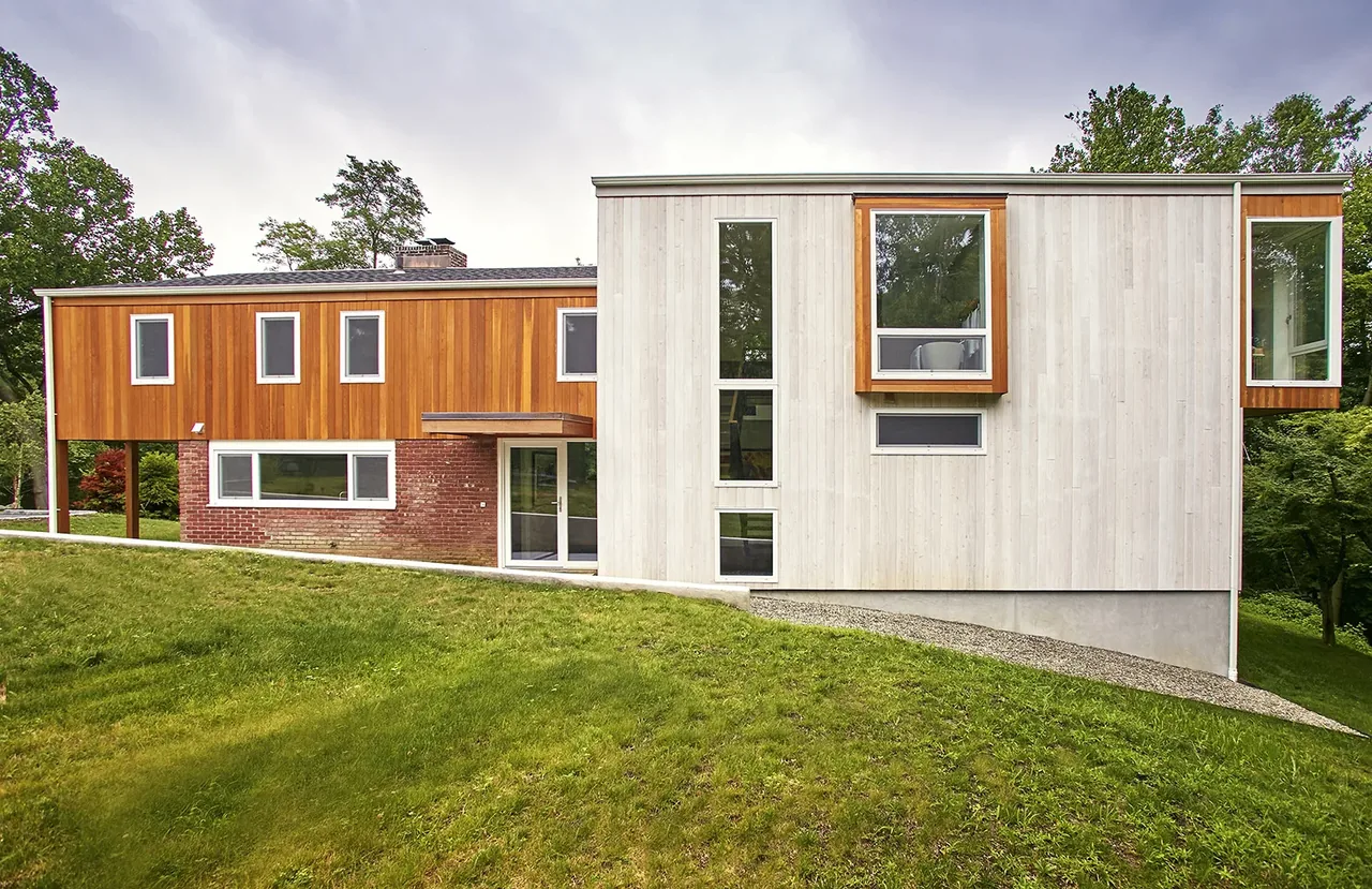 Modern two-story house with wood siding, brick accents, and multiple rectangular windows on a grassy slope.