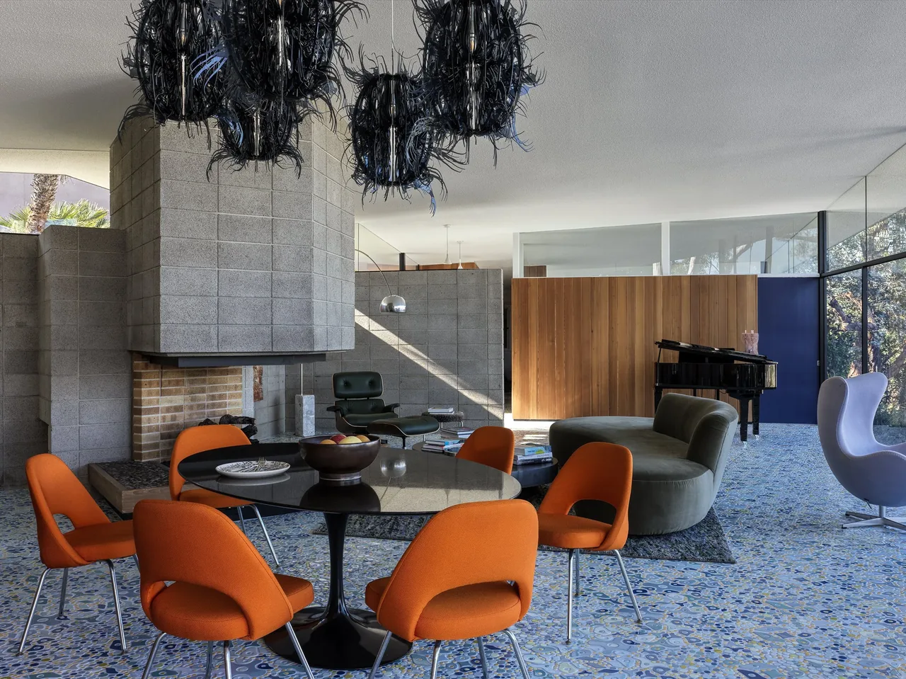 Modern living and dining area with orange chairs, a round black table, a curved green sofa, and a fireplace made of concrete blocks.
