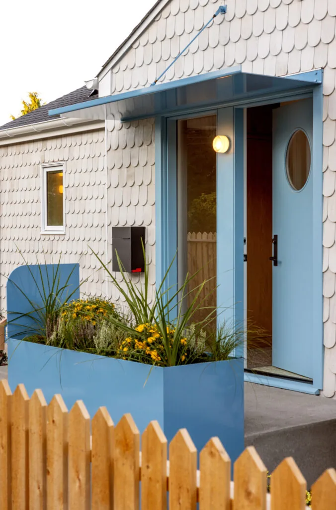 Front entry door painted blue on a house with white shingle siding and a blue metal awning above, flanked by a blue planter with yellow and,