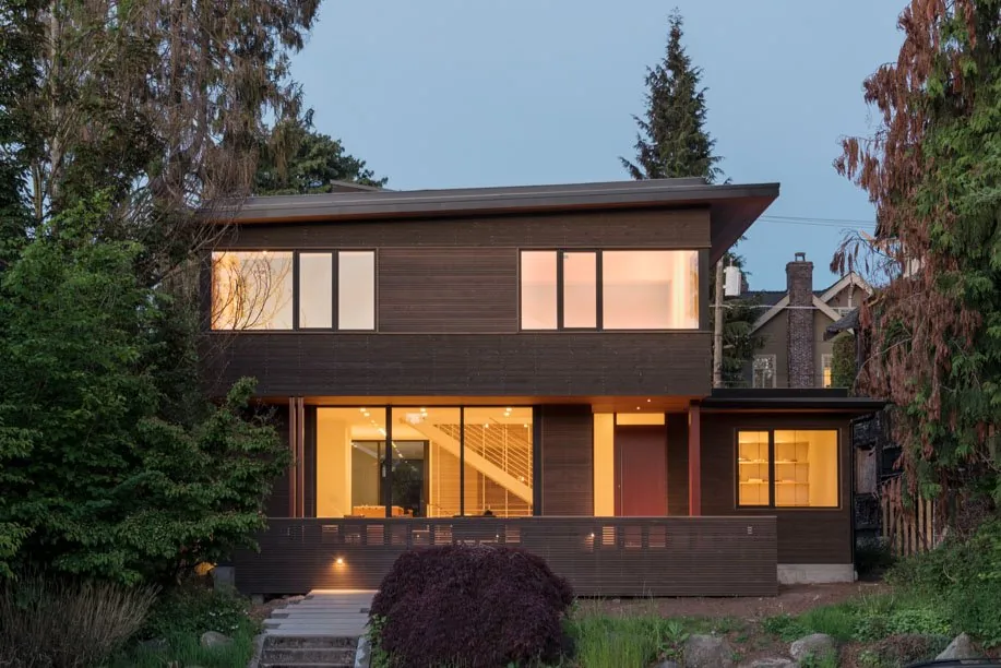 Front view of a two-story modern wood house with large windows lit from inside and a flat roof