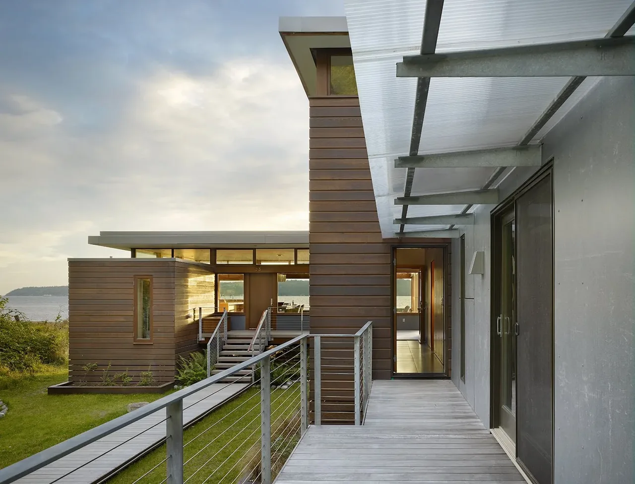 Exterior view of a modern residence with wood siding and a covered walkway overlooking a water body.